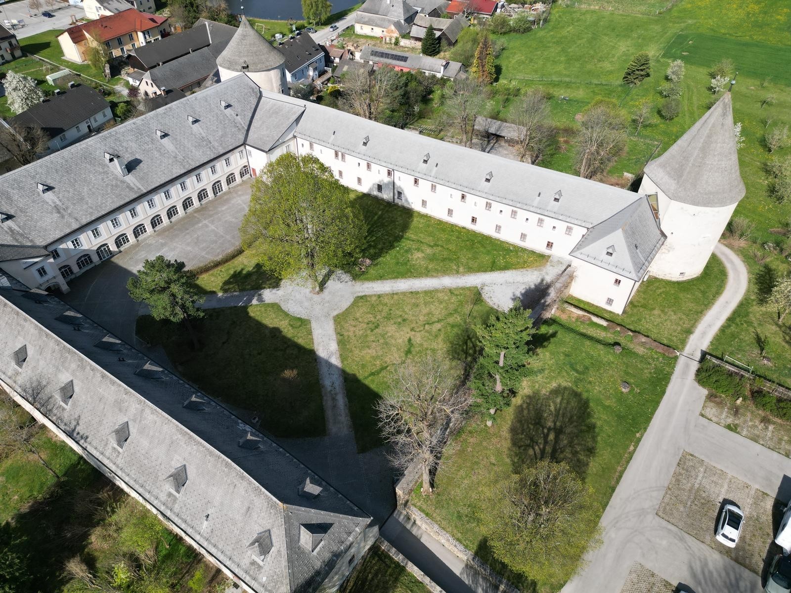 Ein Bild vom Schloss von oben. Es ist zu sehen, dass es ein großer offener Dreikannthof ist. An drei Ecken befinden sich Türme. Im Hof sieht man mehrere Schotterwege sowie einen großen Baum im Zentrum.
